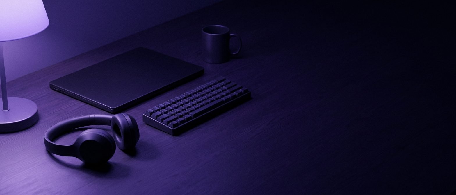 A dark desk lit by a violet lamp, with a closed laptop, headphones, a small keyboard and a mug